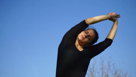 Sportive-young-woman-with-closed-eyes-stretching-against-blue-sky